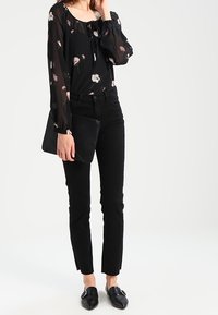 Schwarze Bluse mit floralen Mustern und durchsichtigen langen Ärmeln, kombiniert mit engen schwarzen Hosen und schwarzen Slippern. Hält eine schwarze Clutch.