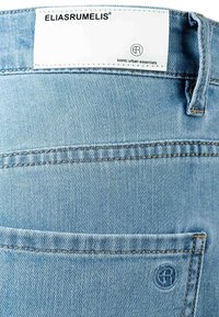 Light blue denim jeans back pocket with stitched logo and white label reading "ELIASRUMELIS iconic urban essentials."