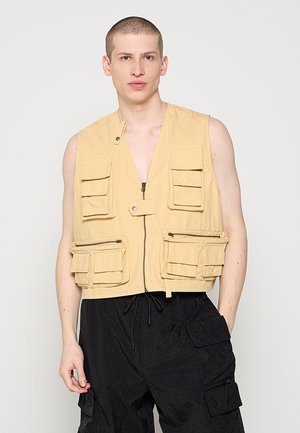 Junger Mann mit kurzem Haar, der eine beige Arbeitweste mit mehreren Taschen und lockere schwarze Cargohosen vor einem einfarbigen Hintergrund trägt.