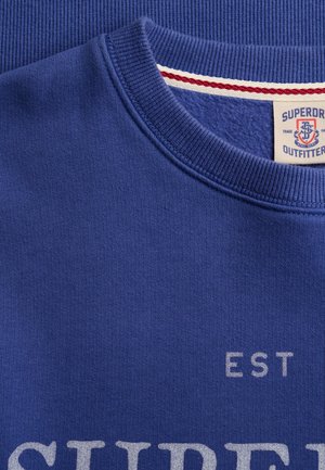 Primer plano de una sudadera azul con cuello redondo acanalado y texto parcial blanco impreso que dice "SUPER" y "EST".