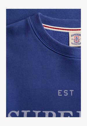 Close-up van een blauwe crewneck-trui met geribbelde kraag en gedeeltelijke witte tekstopdruk "SUPER" en "EST".