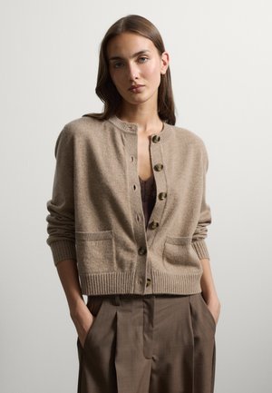 Giovane donna con capelli castani indossa un cardigan beige abbottonato sopra una maglietta marrone, abbinato a pantaloni marroni a vita alta, mani nelle tasche.
