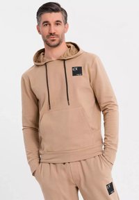 Beige hoodie med framficka, dragsko i huvan och svarta etikett detaljer. Mjukt tyg och åtsittande ärmar, stylad med matchande byxor.