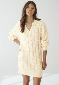 Robe pull en maille couleur crème clair avec un col en V, motif en torsades et poignets côtelés. Texture douce, coupe oversize et longueur mi-cuisse.