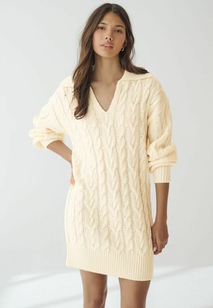 Robe pull en maille couleur crème clair avec un col en V, motif en torsades et poignets côtelés. Texture douce, coupe oversize et longueur mi-cuisse.