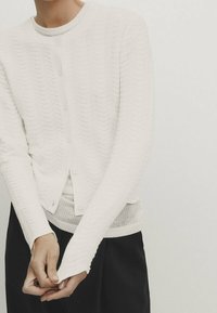 Valkoinen neulecardigan, jossa on aaltoilu kuvio, pyöreä kaula-aukko, nappisulku, pitkät hihat thumbholes-toiminnolla ja pehmeä rakenne.