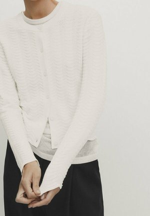 White knit cardigan with a wave pattern, featuring a round neckline, button closure, long sleeves with thumbholes, and a soft texture.