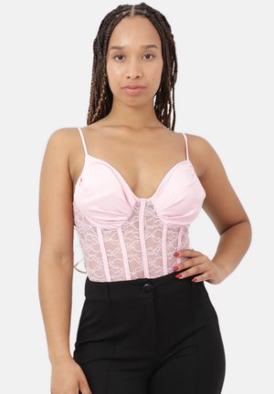 Femme aux longues tresses portant un caraco en dentelle rose à fines bretelles et un pantalon noir taille haute, debout devant un fond uni.