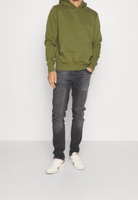 Zelená mikina s kapucí, přední kapsou a malým logem, kombinovaná s tmavými džínami a bílými teniskami. Jednoduchý design, bavlněný materiál, volnější střih.