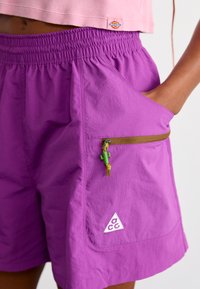 Persona, kas valkā violetus Nike ACG šortus ar elastīgu jostu, sānu rāvējslēdzēja kabatu un zaļu kaktusu rāvējslēdzēja vilkšanu, roka kabatā, piegrieztu rozā topu.