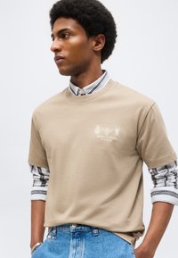 Rövid, göndör hajú férfi, aki bézs pólót visel egy feltűrt csíkos ing fölött és kék farmert egyszerű háttér előtt.