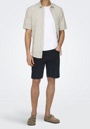 Mann trägt ein beiges, kurzärmeliges Hemd über einem weißen T-Shirt, dunkle Shorts und beigefarbene Slipper, mit einer Hand in der Tasche.