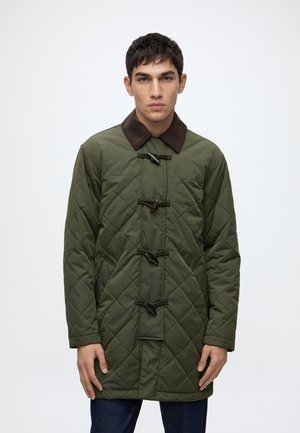 Homme portant un manteau matelassé vert olive avec un col marron et des fermetures à bascule, debout devant un fond clair uni.