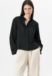 Svart långärmad skjorta med knappslutning och krage, med en texturerad tyg och en bröstficka, kombinerad med krämfärgade veckade byxor.