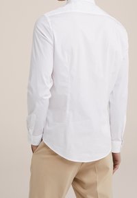 Man in een aansluitend wit long sleeve shirt, gestoken in beige broeken, staand met handen in de zakken, met de rug naar de camera gekeerd.