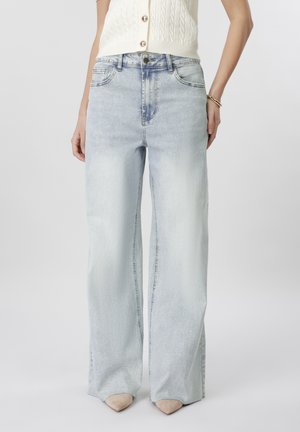 Γυναίκα που φοράει φαρδιά τζιν σε ανοιχτό πλύσιμο, αμάνικο κρεμ πλεκτό τοπ, μπεζ μυτερές γόβες και βραχιόλι στον δεξιό της καρπό.