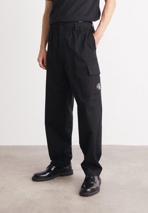 Osoba ubrana w czarne spodnie cargo z logo Calvin Klein oraz czarne skórzane buty sznurowane, stojąca na tle jednolitego jasnego tła.