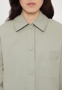Femme portant une chemise vert clair boutonnée avec col et poche poitrine embossée du logo "MAX & Co.", montrée du cou au milieu du torse.