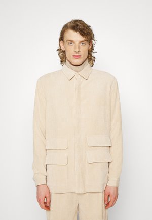 Beige cordfløyelsjakke med knapper foran, krage og to store klaffelommer. Teksturert stoff med vertikale linjer.