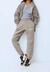 Beige bomullshuvtröja med dragkedja fram och ribbade muddar, matchat med byxor i samma tyg med elastisk midja och sidofickor. Svartvita sneakers.