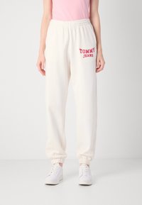 Witte sweatpants hebben een elastische tailleband en boorden bij de enkels, met "TOMMY JEANS" in het rood afgedrukt aan de linkerkant. Gecombineerd met witte sneakers.