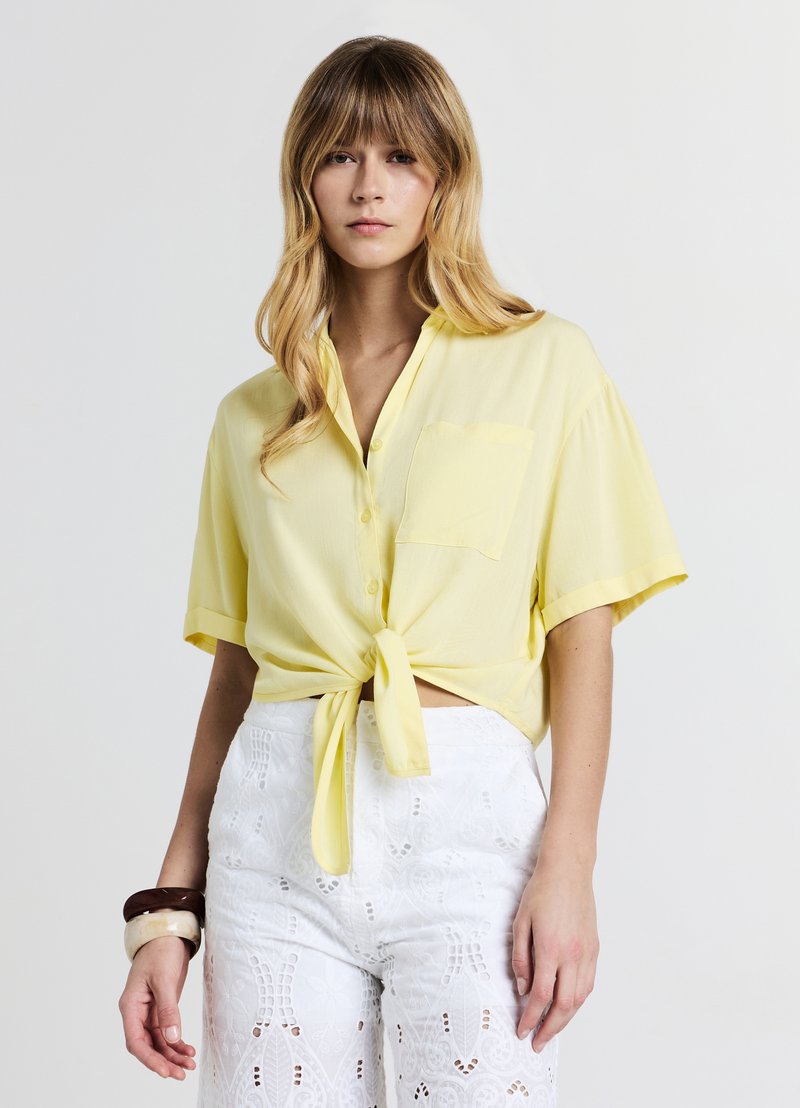 Chemise jaune à manches courtes avec boutons, détail noué à l'avant et poche poitrine, associée à un pantalon blanc brodé taille haute.