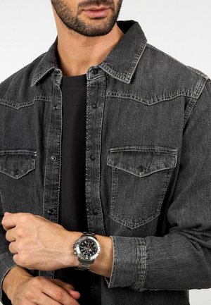 Grijs denim overhemd met knopen aan de voorkant, borstzakken en contrasterend stiksel. Model draagt een metalen zilveren horloge met een zwarte wijzerplaat.