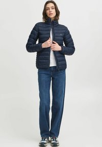 Námornícky modrá vypĺňaná bunda s vysokým golierom, zapínaním na zips a detailmi na bočných švoch, oblečená cez biely top a modré džínsy.