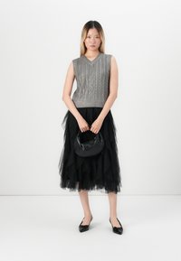 Kvinne står iført en grå ermeløs kabelstrikket genser, lagdelt svart tulle midiskjørt, svarte spissede slingback-sko, og holder en svart håndveske.