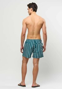 Homme debout de dos, portant un short de bain vert à rayures blanches verticales et des tongs noires sur un fond uni.