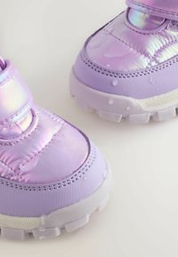 Lila wasserdichte Schuhe mit glänzender Oberfläche, Klettverschluss, strukturiertem Sohlenprofil und sichtbaren Wassertropfen auf der Oberfläche.