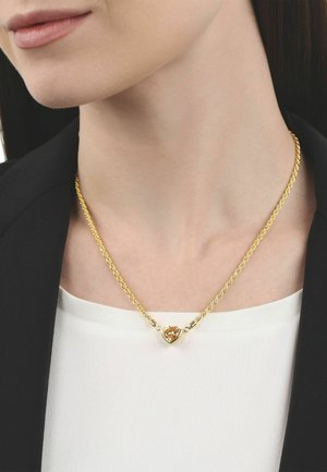 Collar de cadena torcida de oro con un colgante de gema ámbar en forma de corazón, llevado sobre una camiseta blanca y una chaqueta negra.