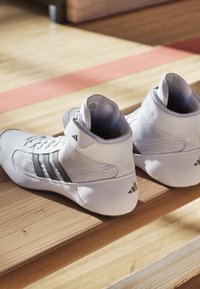 White high-top athletic shoes made of mesh and synthetic material, featuring three black stripes and a padded ankle support, placed on a wooden surface.