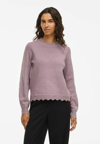 Femme aux cheveux bouclés et foncés portant un pull en maille lavande avec des bords en festons et un pantalon noir, se tenant contre un fond blanc uni.