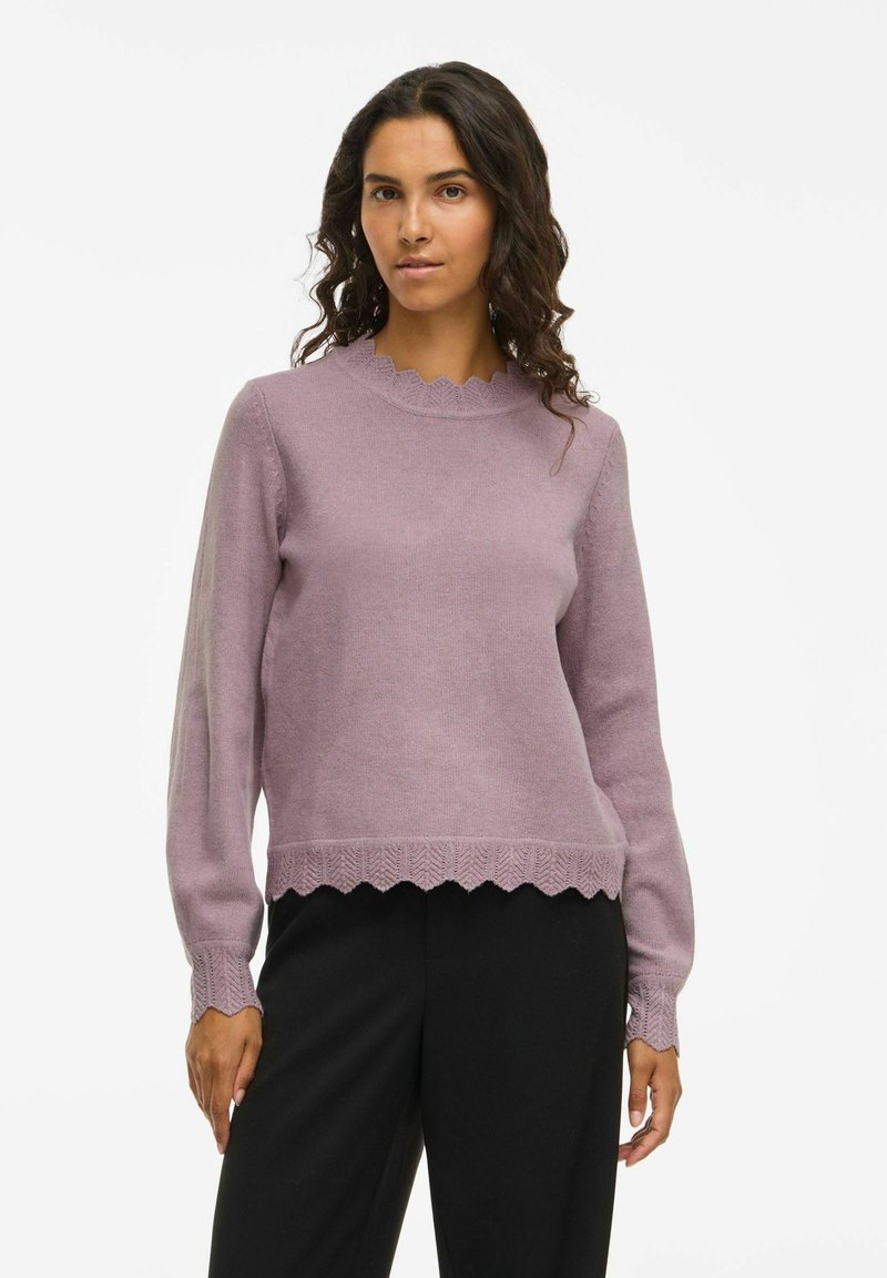 Femme aux cheveux bouclés et foncés portant un pull en maille lavande avec des bords en festons et un pantalon noir, se tenant contre un fond blanc uni.