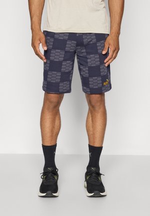 Mand iført marineblå Puma-shorts med ternet logo mønster, sorte Puma-strømper og sorte løbesko, der står mod en ensfarvet baggrund.