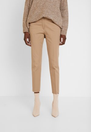 Person, die beige Slim-Fit-Hosen, hellbeige Stiefeletten und einen braunen Strickpullover trägt und vor einem schlichten weißen Hintergrund steht.