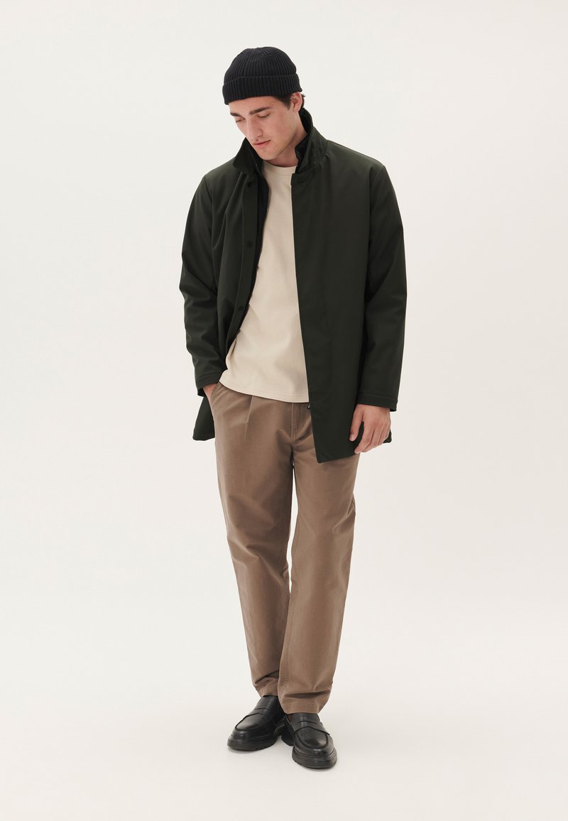 Chaqueta verde oscuro con cuello alto, camisa beige, pantalones marrón claro y zapatos negros; acabado de tejido suave con un ajuste relajado.
