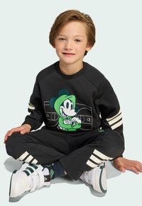 Čierna mikina so zelenou grafikou Mickey Mouse, čierne tepláky s bielymi pruhmi a biele topánky so zelenými akcentmi; hladká textúra.