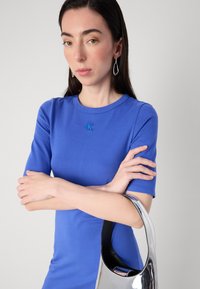 Mladá žena s dlouhými, tmavými vlasy si překřížila ruce, má na sobě modrou košili a stříbrné náušnice a drží kovovou kabelku.