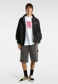 Svart vindjacka med gröna ärmadjusteringar, vit grafisk t-shirt med blommönster, grå denimshorts och svarta sneakers med flamamönster.