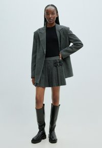 Grau karierter Blazer, kombiniert mit einem schwarzen Oberteil und einem plissierten Rock mit zwei schwarzen Schnallenakzenten. Abgerundet mit schwarzen kniehohen Stiefeln.