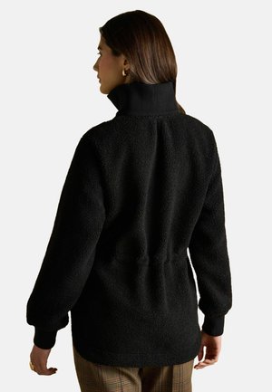 Woman with brown hair wearing gold hoop earrings, a black textured fleece jacket, and plaid pants, viewed from behind on a plain background.