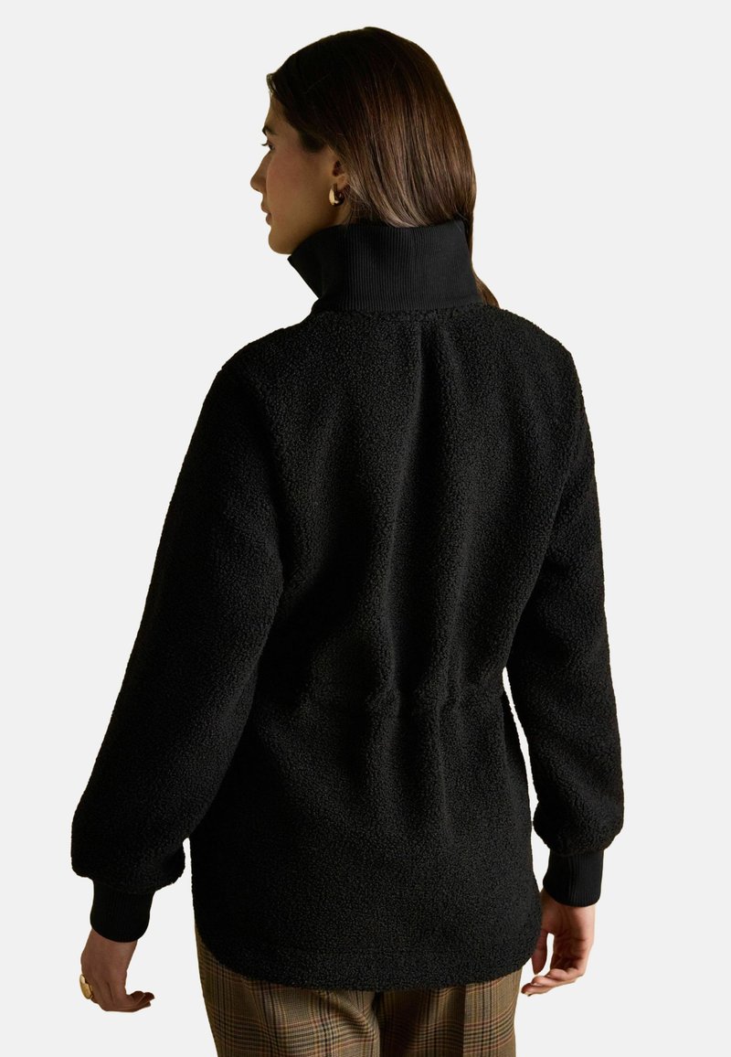 Kobieta z brązowymi włosami, nosząca złote kolczyki w kształcie kół, czarną teksturowaną kurtkę polarową i spodnie w kratę, widoczna z tyłu na jednolitym tle.