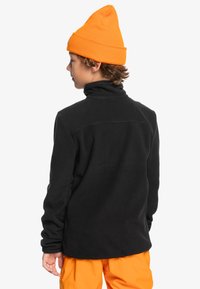 Schwarzer Fleece-Pullover mit hohem Kragen, getragen unter einer orangefarbenen Mütze. Hat lange Ärmel und eine figurbetonte Silhouette mit Flachnähte.