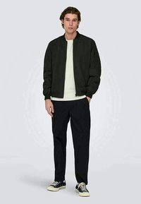 Black bomber jacket with a zip front, ribbed cuffs, and side pockets, worn over a light shirt and paired with dark pants and sneakers.