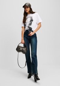 White graphic t-shirt with black design, blue flared jeans, black heeled boots, and a grey cap. Holding a black handbag with a logo.
