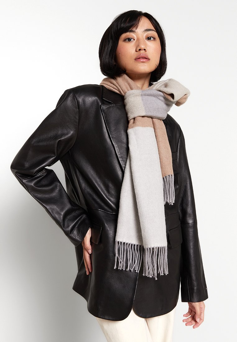 Mehrfarbiger Schal in den Farben Beige, Grau und Creme, aus einem weichen Material mit franjierten Kanten, getragen über einer schwarzen Lederjacke.