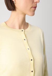 Cardigan jaune clair en matière tricotée douce, doté d'un col rond et de cinq boutons métalliques argentés sur le devant.