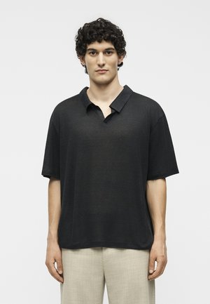 Άνδρας με σγουρά σκούρα μαλλιά, που φοράει μαύρο κοντομάνικο πουκάμισο με γιακά και ανοιχτό μπεζ παντελόνι, όρθιος μπροστά σε ένα απλό ανοιχτό φόντο.